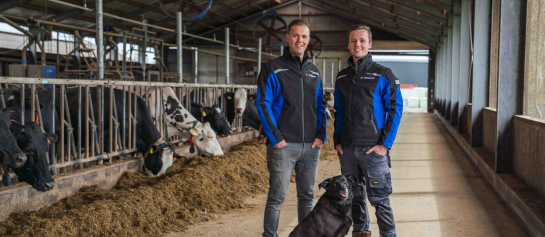 Melkveehouders van Houten met hun hond op de voergang en koeien aan het voerhek